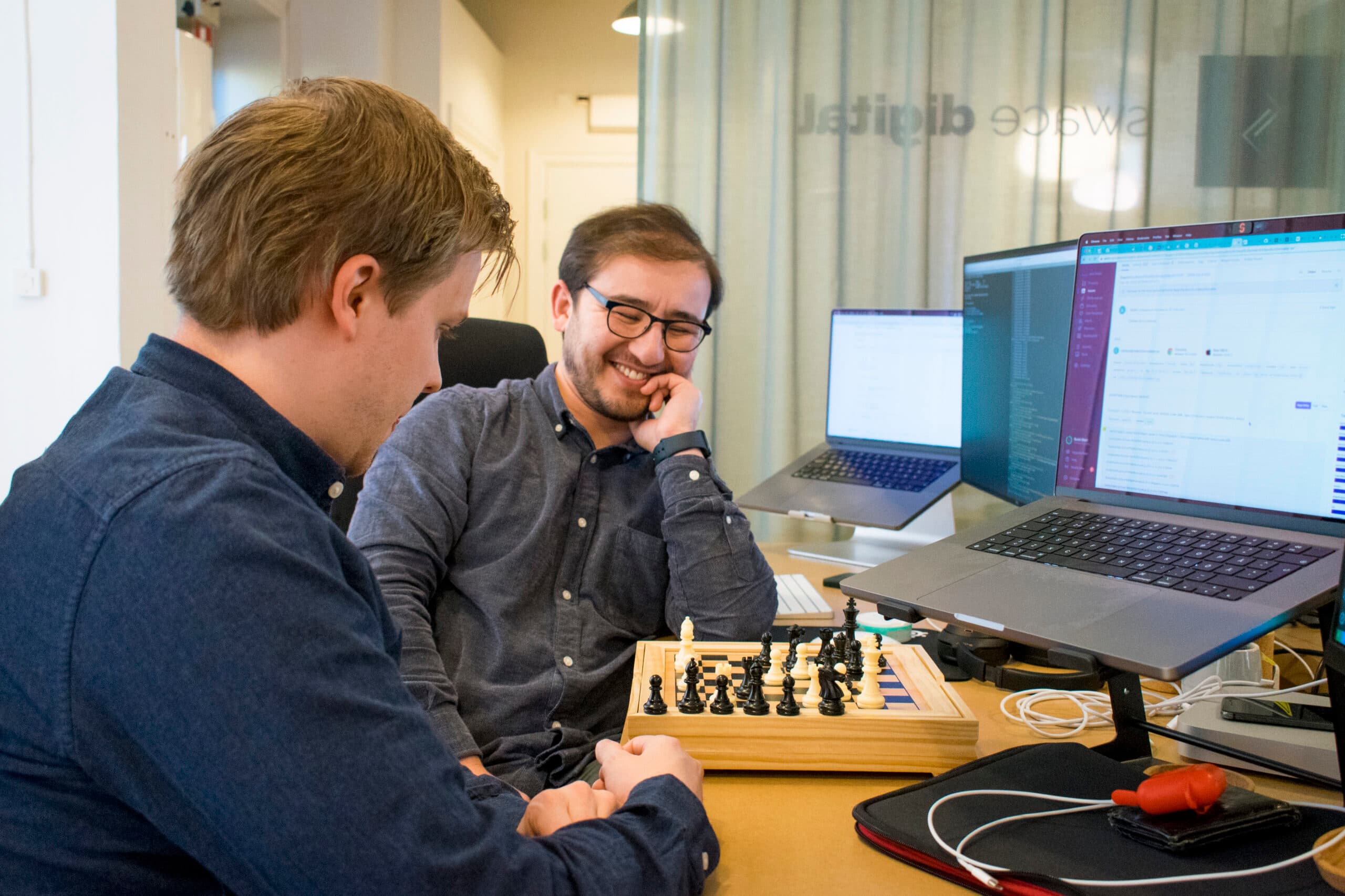 Two colleagues sitting at a desk in the Swace office, smiling while playing chess during a break.