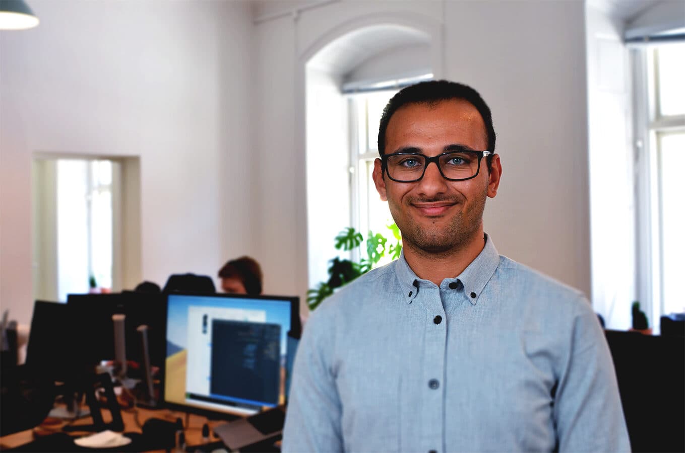 A Swace colleague standing and smiling at the office, with desks and computers in the background.