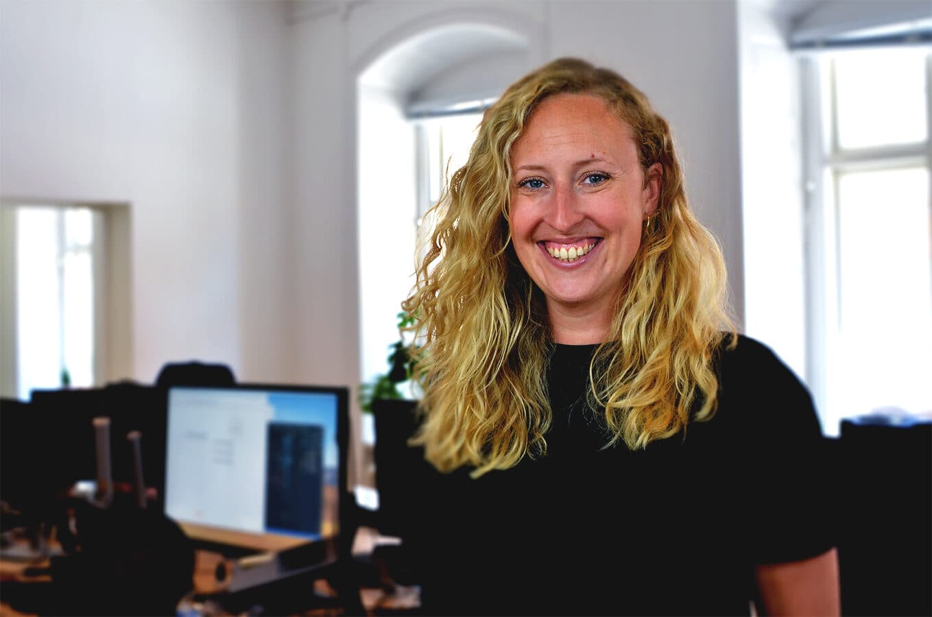 A Swace colleague smiling at the office, standing by a desk with computers in the background.
