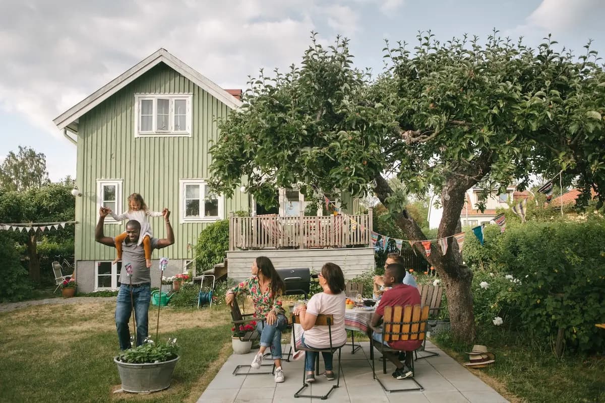 People gathered for a garden meal outside a green wooden house, with a child sitting on an adult’s shoulders