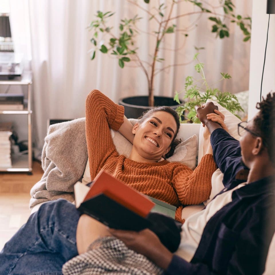 Two people relaxing together on a sofa at home, smiling and holding hands with books in their laps