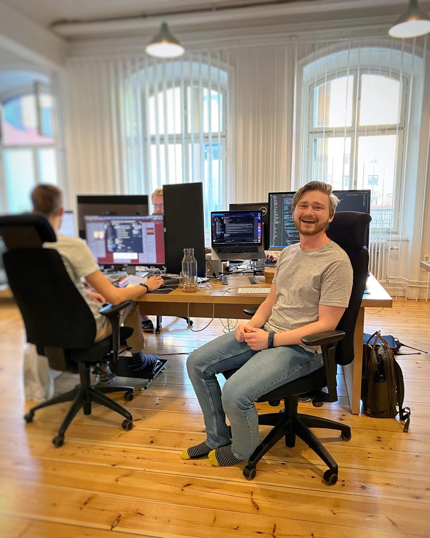 Two colleagues working at their desks in the office. One turns toward the camera and smiles.