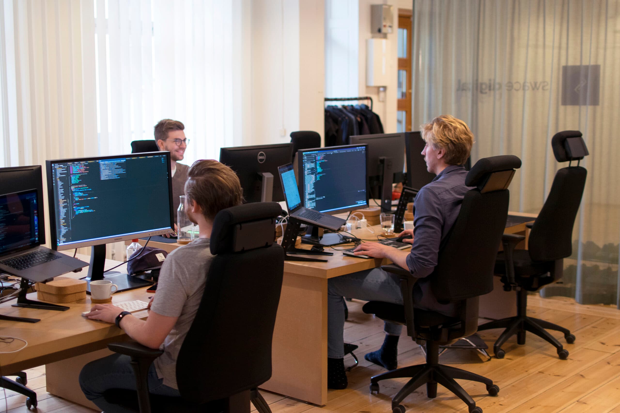 Several colleagues working at their computers in the office, talking while coding.