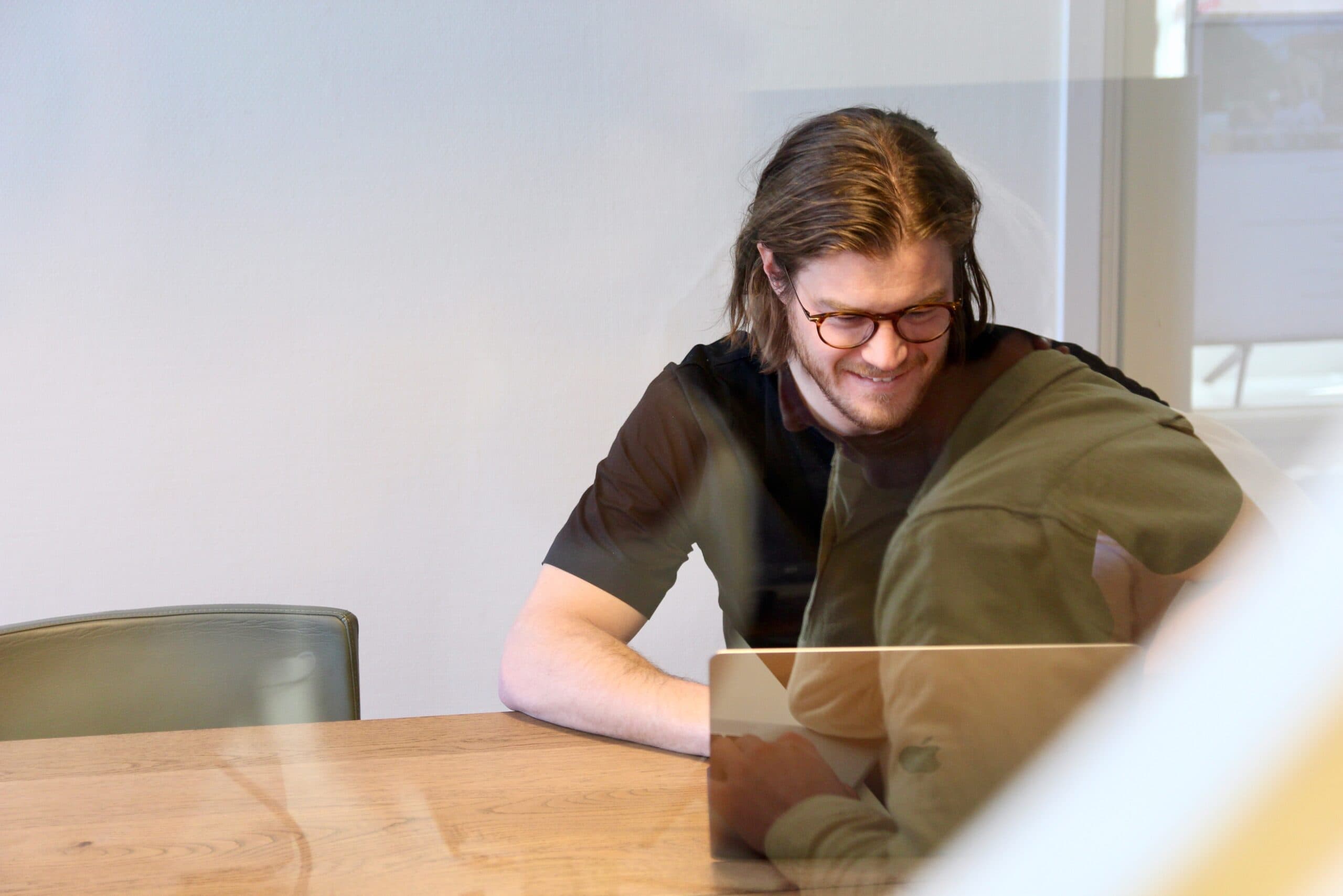 A colleague sits at a meeting table with a laptop, smiling during a conversation.