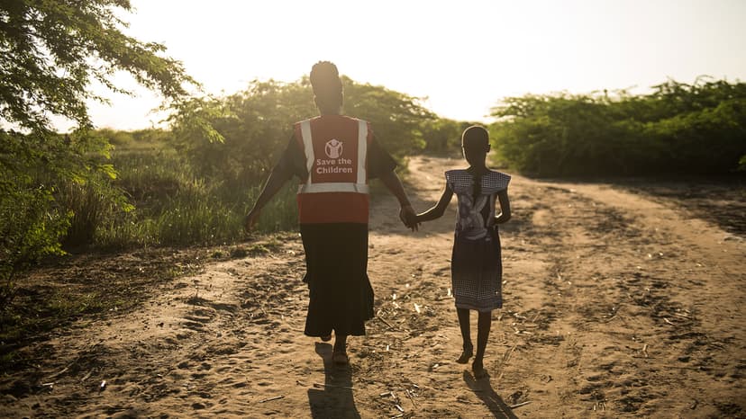 Save the Children worker walking hand in hand with a child along a dirt path in the evening light