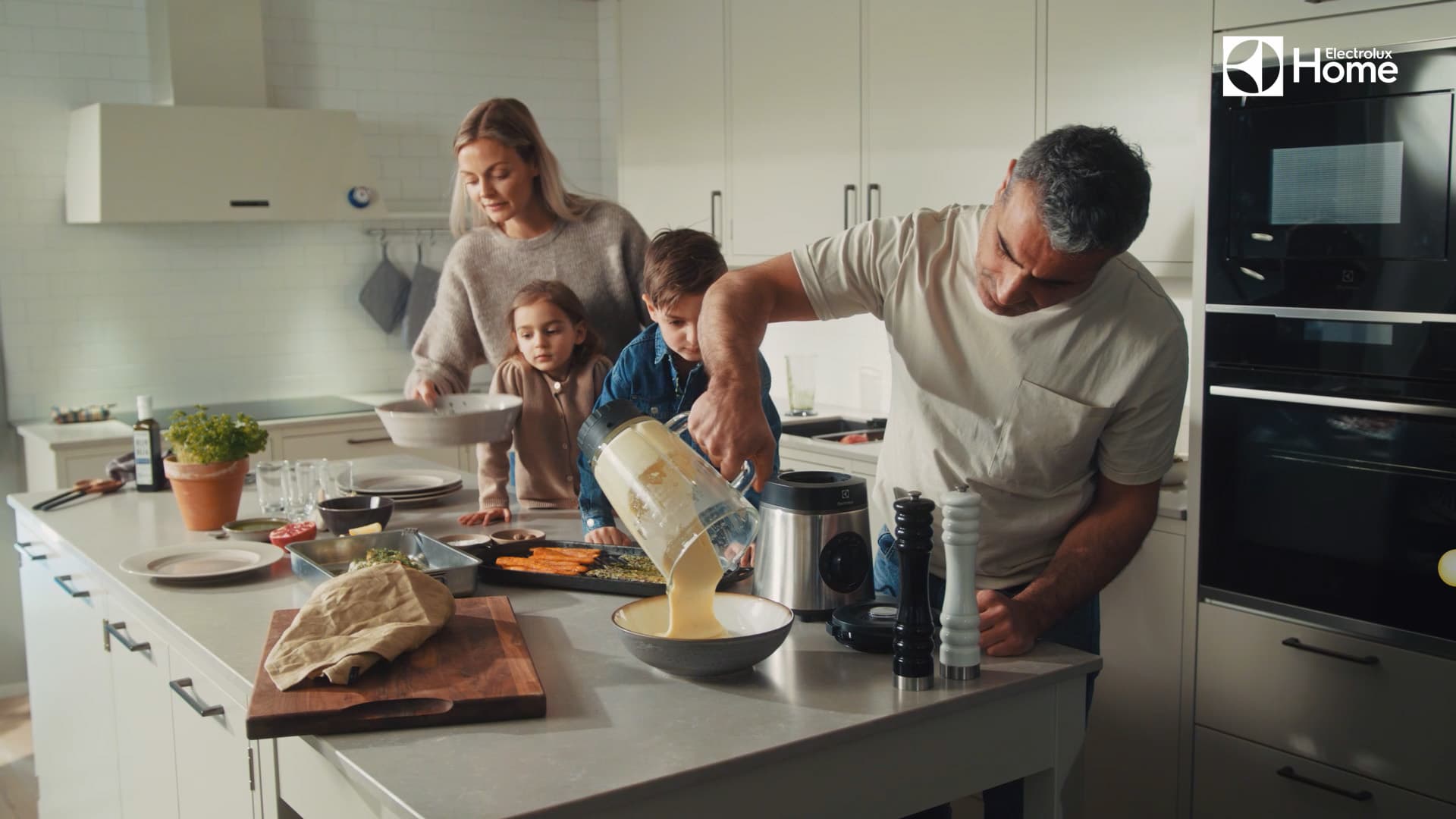 A family cooking together in a modern kitchen. A man pours batter from a blender while two children and a woman assist at the kitchen island. Electrolux appliances are visible in the background.